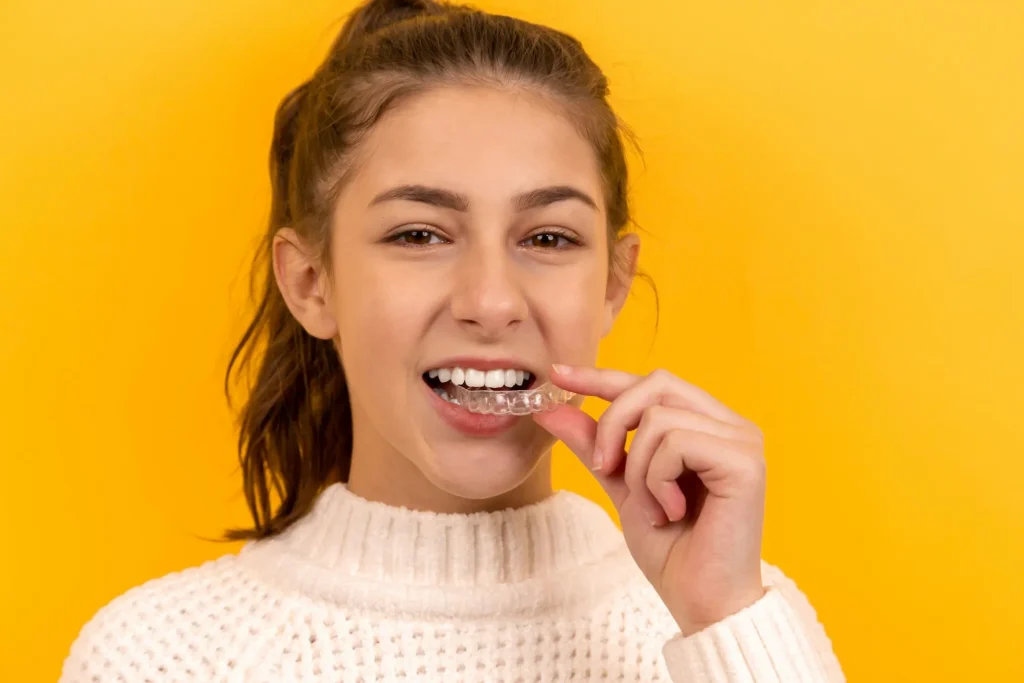 Girl holding a clear mouthguard, smiling against a yellow background, illustrating the importance of custom mouthguards for dental protection in Deer Park, TX.